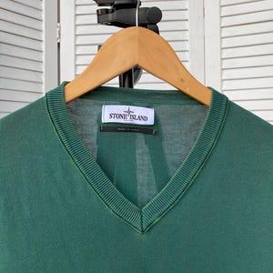 Pu&ograve; includere: Un maglione verde scuro a collo a V con un'etichetta logo Stone Island. L'etichetta &egrave; bianca con testo nero e un logo a bussola.