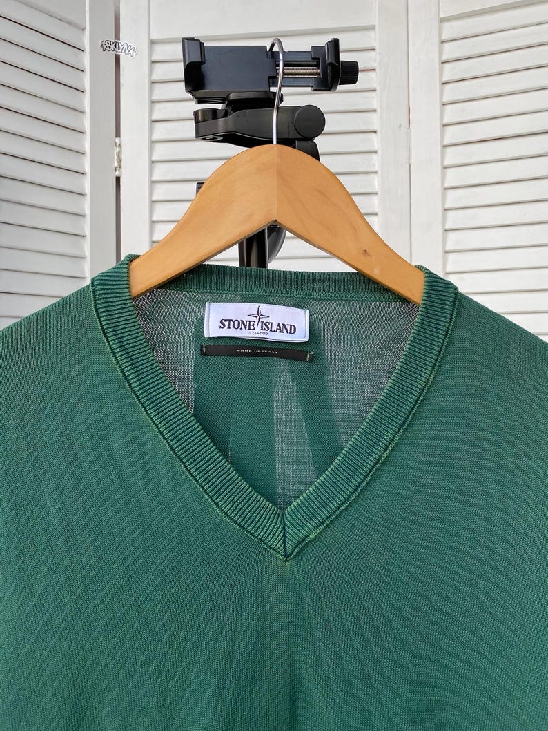 Pu&ograve; includere: Un maglione verde scuro a collo a V con un'etichetta logo Stone Island. L'etichetta &egrave; bianca con testo nero e un logo a bussola.