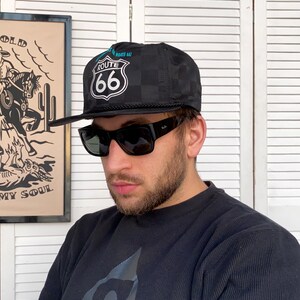 Può includere: Un cappello da baseball a scacchi neri e bianchi con un logo Route 66 blu e bianco. Il cappello ha una visiera nera e un cinturino nero.