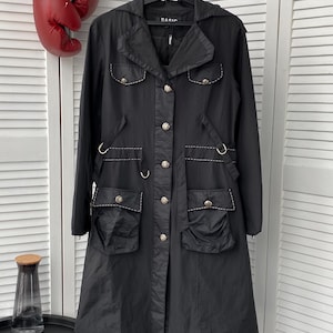 Puede incluir: Una chaqueta negra de manga larga con cierre de botones, cuello y varios bolsillos. La chaqueta tiene botones plateados y detalles de costura.