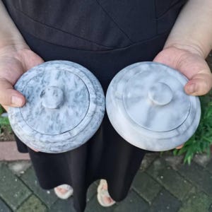 May include: Two round, grey marble containers with lids are held up. Each lid has a small, circular knob in the centre. The marble has a natural, swirled pattern of grey and white. The containers appear to be small, possibly for storing jewellery or other small items.