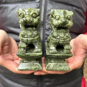 May include: Two green jade lion statues. Each statue is carved with intricate details, including a fierce expression and ornate detailing. The lions are mounted on rectangular bases. The statues are held in a person's hands.