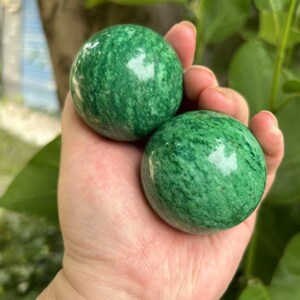 Op de afbeelding: Twee gepolijste groene bollen met een gevlekt patroon worden in een hand gehouden. De bollen hebben een glad, reflecterend oppervlak en een levendige groene kleur, met donkerdere groene adering.