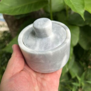 May include: A small, cylindrical container crafted from light gray and white marbled stone. The vessel features a smooth, polished surface with natural veining patterns and includes a matching circular lid with a central knob handle on top.