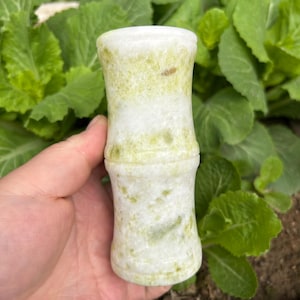 May include: A bamboo-shaped, white and green stone sculpture. The cylindrical object has a textured surface with green veining and is held in a hand against a backdrop of green leaves. The sculpture is a decorative item.