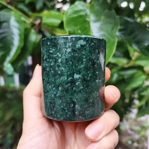 Pode incluir: Uma caneca cilíndrica de pedra verde escura com um padrão salpicado. A caneca é segurada em uma mão, com um fundo desfocado de folhagem verde. A superfície da caneca tem uma aparência polida, mostrando as variações naturais da pedra.