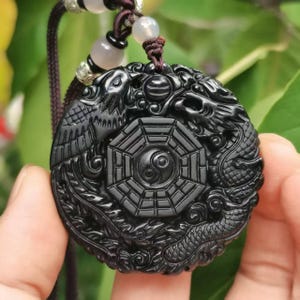 May include: A black, circular pendant carved with a dragon and bird motif, framing a Yin Yang symbol. It hangs from a brown cord with grey and white beads. The pendant's detailed design is highlighted in the dark stone.