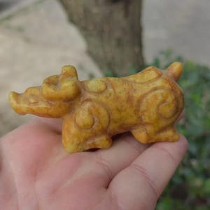 May include: A small, carved figurine of an animal, possibly a pig or ox, in shades of yellow and brown. The sculpture has rounded features and decorative swirl patterns. The figurine is held in a person's hand.