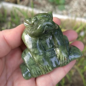 May include: A carved green stone figurine of a bear-like creature. The sculpture has detailed features, including a defined face, paws, and body. The stone has a polished, glossy finish, and the color varies from light to dark green.