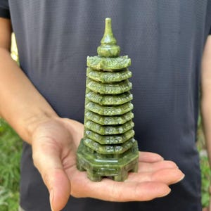 Puede incluir: Escultura de pagoda de jade verde con múltiples niveles y una parte superior puntiaguda. La pagoda se sostiene en una mano, destacando su diseño detallado y color. El jade tiene un acabado pulido. La escultura mide unos 10 cm de altura.