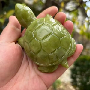 Könnte beinhalten: Eine jadegrüne Schildkrötenfigur mit detailliertem Panzer und Gliedmaßen. Die Schildkröte ist aus einem massiven Steinblock geschnitzt und wird in einer Hand gehalten. Die Figur hat eine glatte, polierte Oberfläche und ist ein dekorativer Gegenstand.