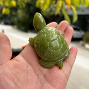 May include: A small, jade-green turtle figurine. The turtle is carved with detailed features, including a textured shell and visible limbs. The figurine is a solid, polished green colour, and is held in a hand.