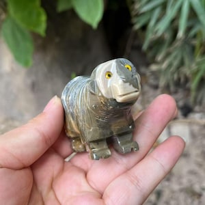Puede incluir: Una figura de oso de piedra tallada, sostenida en una mano. El oso es principalmente gris y marrón con ojos amarillos. La escultura tiene un acabado liso y pulido y está detallada con líneas talladas.