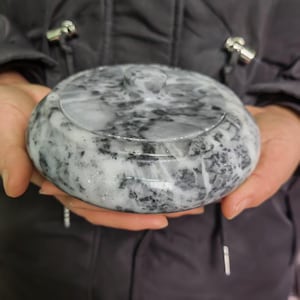 Può includere: Un contenitore rotondo con coperchio, con un motivo marmorizzato grigio e bianco. Il coperchio ha un piccolo pomello arrotondato. Il contenitore è tenuto con entrambe le mani, mostrando la sua superficie liscia e lucida. Il design suggerisce un oggetto decorativo o di stoccaggio.