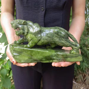 Op de afbeelding: Een gebeeldhouwde groene stenen tijgersculptuur. De tijger wordt afgebeeld in een hurkende positie met open mond, gemonteerd op een rechthoekige basis. De sculptuur wordt voor een donkere achtergrond gehouden.