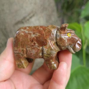 May include: A small, hand-carved stone figurine of a rhinoceros. The sculpture features a mottled brown and tan colour palette with a polished finish. It has a distinct, bright yellow circular eye inlay on the side of its head and carved details.