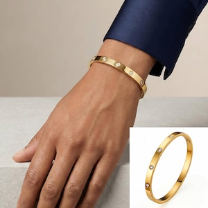 Pode incluir: Pulseira de bracelete dourada com seis pedras preciosas redondas e transparentes, espaçadas na parte superior. A pulseira é mostrada em um pulso, com uma manga azul marinho ao fundo. A pulseira também é mostrada separadamente.