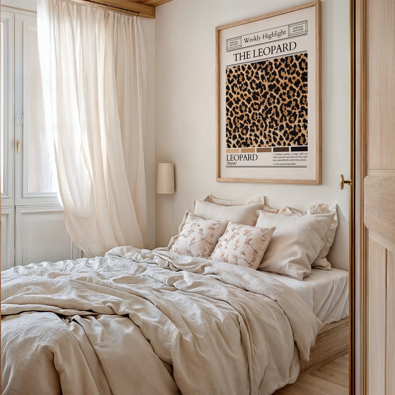 May include: A bedroom with a white bed with a beige linen duvet and pillows. The bed is next to a window with white curtains. A framed print with a leopard print pattern and the text "The Leopard" is on the wall.