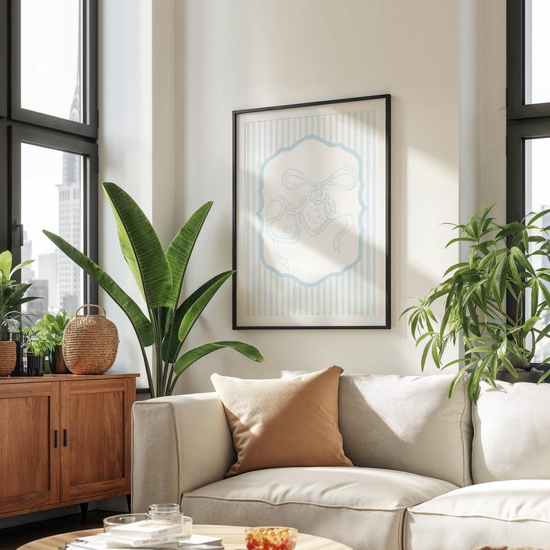 May include: A framed art print featuring a light blue and white design. The artwork is in a black frame and hangs on a white wall above a beige sofa. The room also includes a wooden cabinet, plants, and a window.