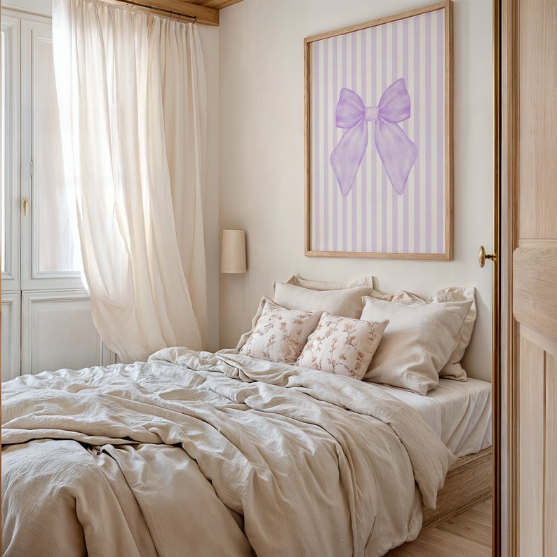 May include: A bedroom with a white bed with a beige linen duvet and pillows. The bed is next to a window with white curtains. There is a framed print on the wall with a purple bow on a striped background.