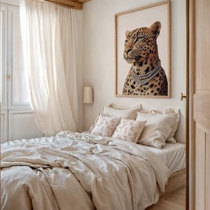 May include: A beige linen bed with a white and pink floral patterned pillow and a white pillow. A framed print of a leopard wearing a diamond necklace hangs on the wall above the bed.