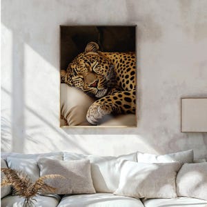 Pu&ograve; includere: Stampa artistica incorniciata raffigurante un leopardo addormentato. Il leopardo ha un mantello beige con macchie nere e riposa su un cuscino beige. L'opera d'arte &egrave; esposta su una parete bianca sopra un divano bianco.