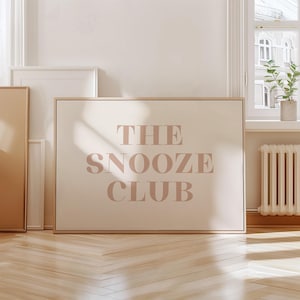 Puede incluir: Un cuadro enmarcado con el texto "THE SNOOZE CLUB" en un tono rosa suave. La obra de arte está sobre un fondo color crema y enmarcada en madera clara. El cuadro se exhibe en una habitación con una ventana y un radiador.