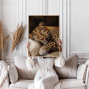 Pu&ograve; includere: Una stampa incorniciata raffigurante un leopardo addormentato con un mantello maculato marrone e nero, appoggiato su un cuscino color crema. L'opera d'arte &egrave; tenuta da una persona che indossa un vestito bianco, seduta su un divano beige.