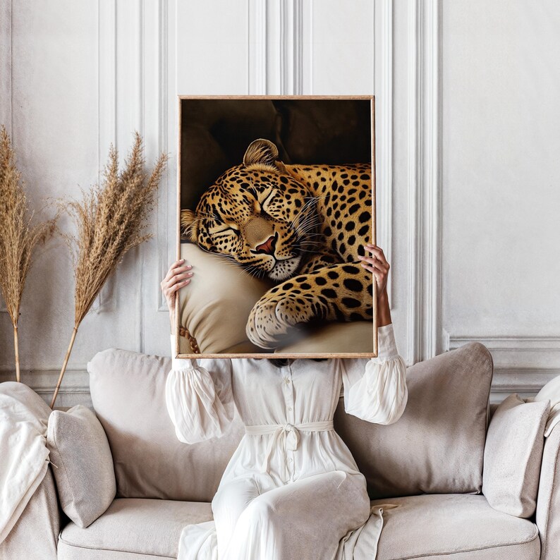 Pu&ograve; includere: Una stampa incorniciata raffigurante un leopardo addormentato con un mantello maculato marrone e nero, appoggiato su un cuscino color crema. L'opera d'arte &egrave; tenuta da una persona che indossa un vestito bianco, seduta su un divano beige.