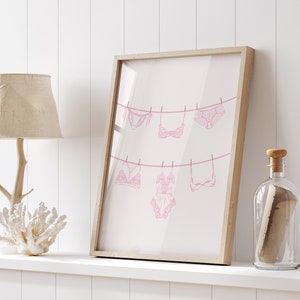May include: A framed print of pink lingerie hanging on a clothesline. The print is on a white background and is framed in a light wood frame.