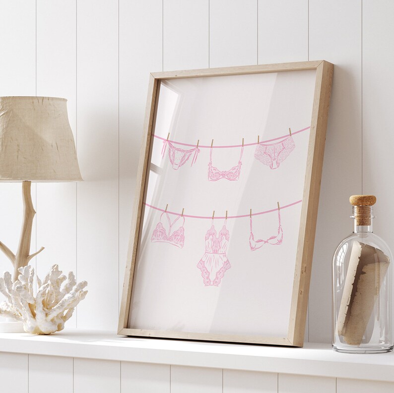 May include: A framed print of pink lingerie hanging on a clothesline. The print is on a white background and is framed in a light wood frame.