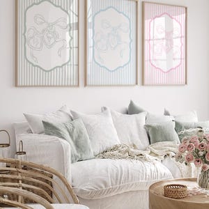 May include: Three framed prints with a bow and ribbon design. The prints have a light blue, light blue, and pink border and background. The prints are displayed above a white sofa with decorative pillows and a wooden coffee table.