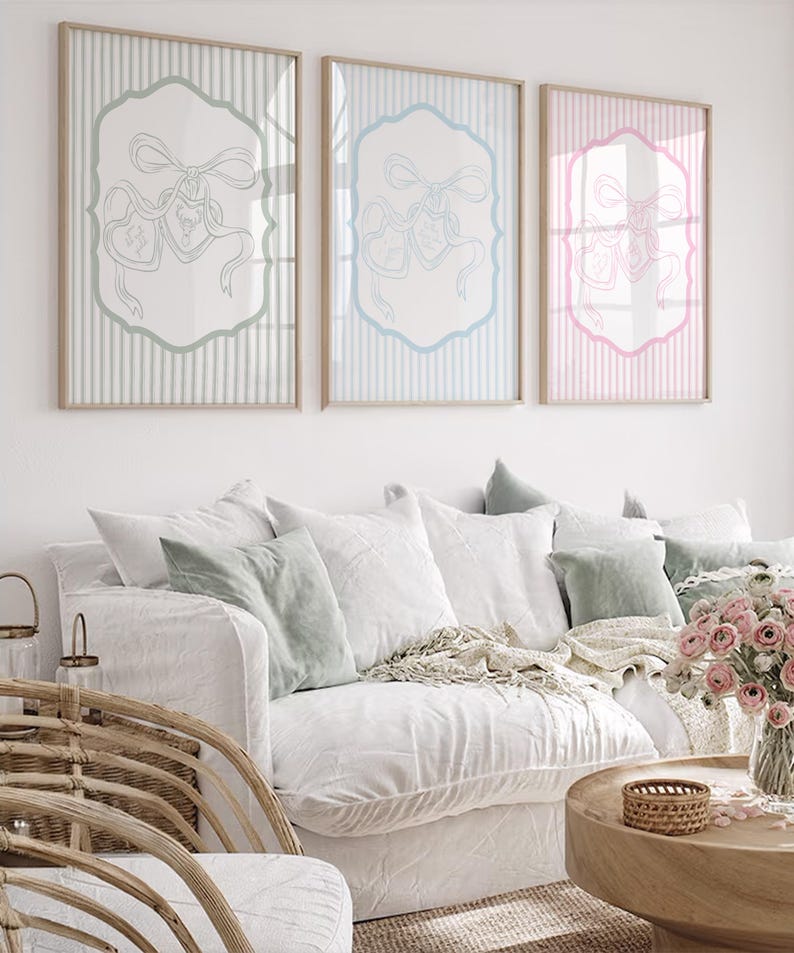 May include: Three framed prints with a bow and ribbon design. The prints have a light blue, light blue, and pink border and background. The prints are displayed above a white sofa with decorative pillows and a wooden coffee table.