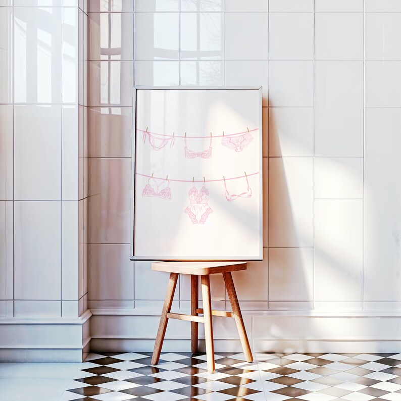 May include: A framed print of pink lingerie hanging on a clothesline. The print is in a white frame and is hanging on a white tiled wall.