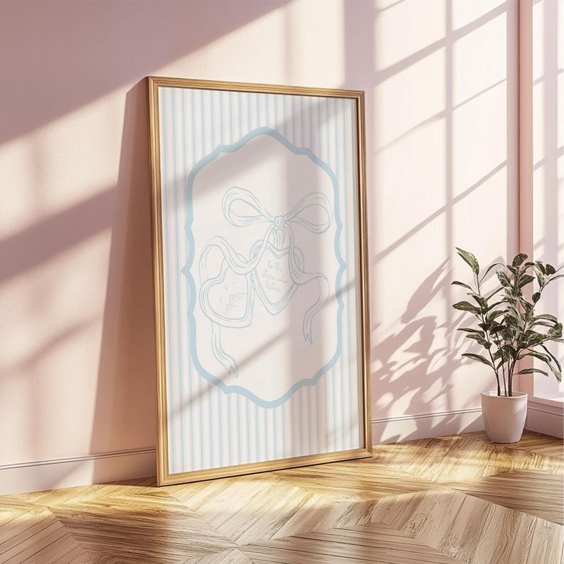 May include: A framed artwork with a gold border, leaning against a pink wall. The artwork features a light blue and white striped background with a light blue decorative frame. Inside the frame are two heart-shaped ribbons with text.