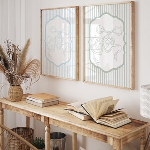 May include: Two framed prints with light blue and white striped backgrounds and a light blue decorative design. The prints are displayed above a wooden table with books and a woven basket. The room has a neutral color palette.