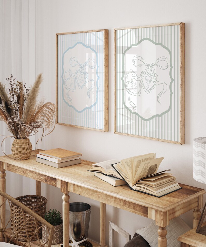 May include: Two framed prints with light blue and white striped backgrounds and a light blue decorative design. The prints are displayed above a wooden table with books and a woven basket. The room has a neutral color palette.