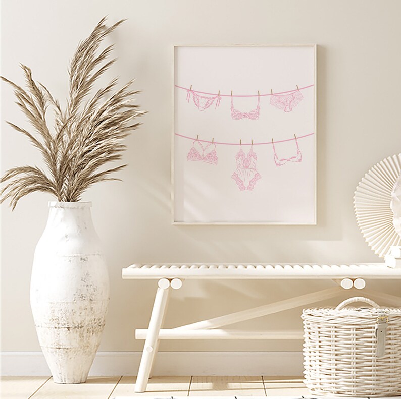 May include: A pink and white print of lingerie hanging on a clothesline. The print is framed and hanging on a white wall.