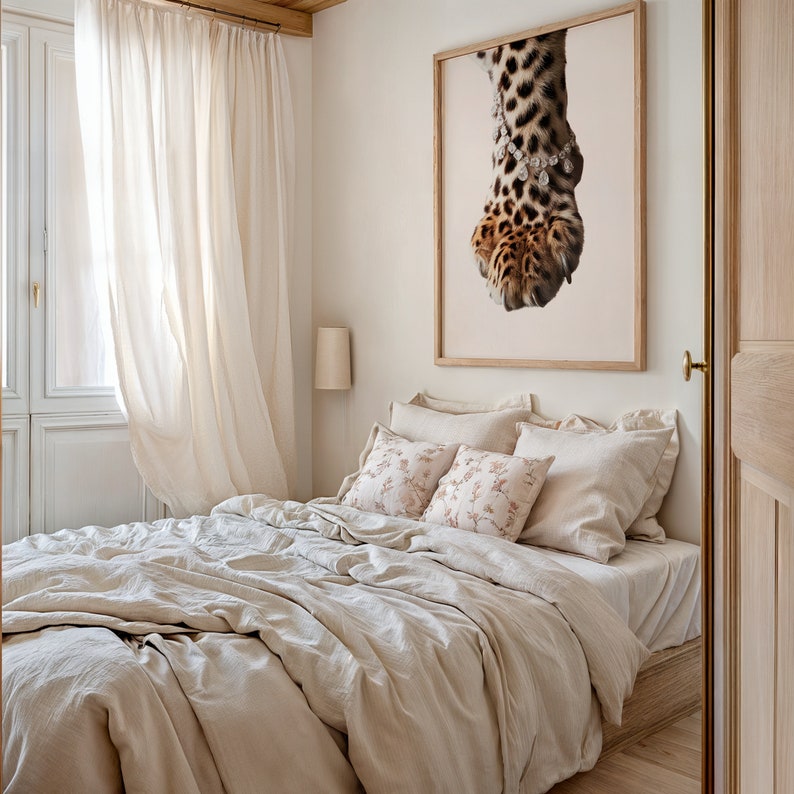 May include: A white linen bed with a beige duvet cover and two decorative pillows with floral patterns. A framed print of a leopard paw with a diamond necklace hangs on the wall above the bed.