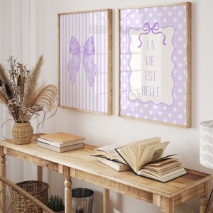 May include: Two framed prints with a purple and white polka dot pattern and a purple and white striped pattern. The print with the polka dot pattern has a white frame with a purple bow and the text "La Vie Est Belle". The print with the striped pattern has a white frame with a purple bow.