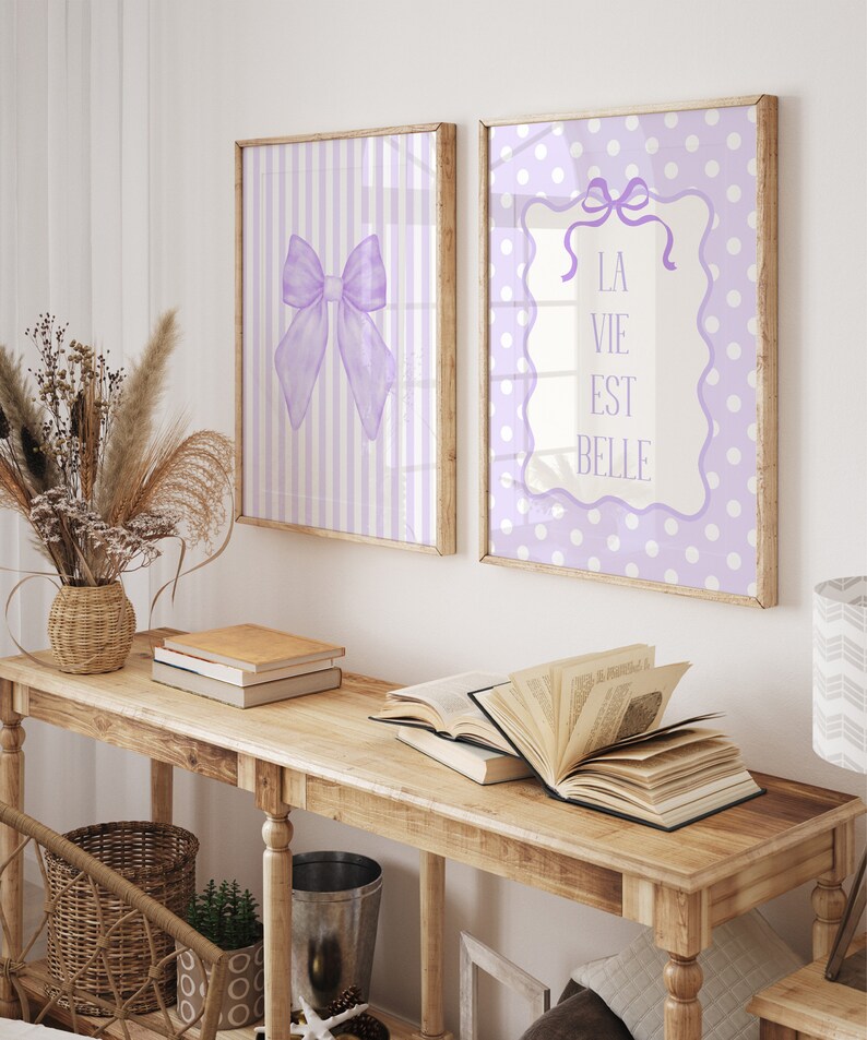 May include: Two framed prints with a purple and white polka dot pattern and a purple and white striped pattern. The print with the polka dot pattern has a white frame with a purple bow and the text "La Vie Est Belle". The print with the striped pattern has a white frame with a purple bow.