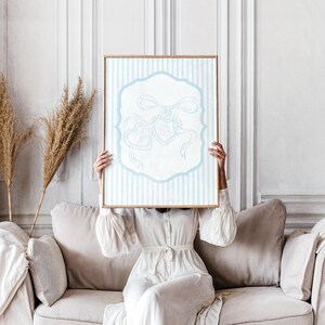 May include: A framed artwork featuring a light blue and white striped background with a central white design. The design includes a light blue ribbon and heart-shaped elements. The artwork is held by a person wearing a white dress.