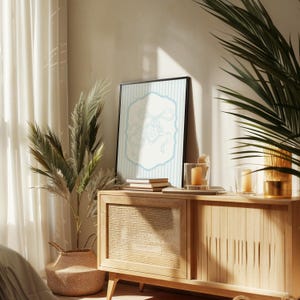 May include: A framed art print featuring a light blue illustration on a white background with a light blue border, set against a striped backdrop. The artwork is displayed on a wooden cabinet, complemented by candles and decorative plants.