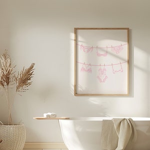 May include: A framed print of pink lingerie hanging on a clothesline. The print is in a light wood frame and is hanging on a white wall.