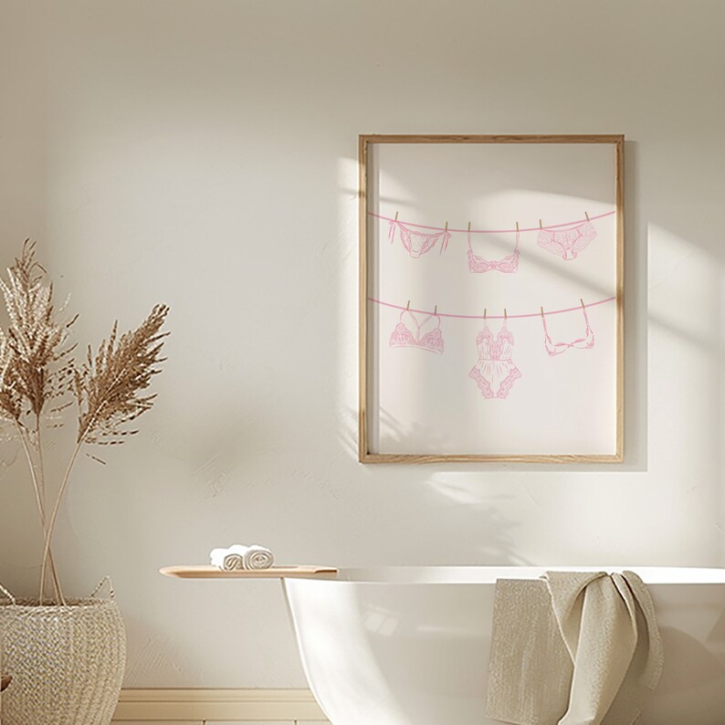 May include: A framed print of pink lingerie hanging on a clothesline. The print is in a light wood frame and is hanging on a white wall.