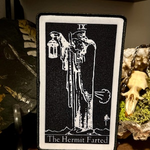 May include: A black and white embroidered patch with a tarot card design. The card depicts the Hermit, a figure with a lantern and a staff, with a cloud of gas coming from behind them. The text on the card reads "The Hermit Farted".