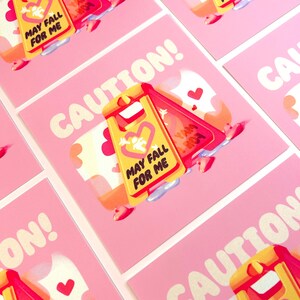 May include: A pink and yellow caution sign with the text "May Fall For Me" and a heart. The sign is on a pink background with hearts and the word "CAUTION!" in white.