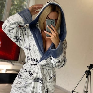 May include: A white hooded robe with a blue trim and a pattern of black text and designs. The robe is made of a soft, plush material and has a tie closure. The robe is being worn.