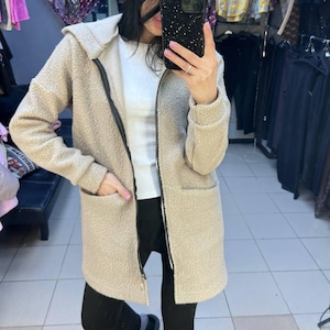Può includere: Una giacca beige a maniche lunghe con cappuccio, realizzata in un materiale testurizzato simile al pile. La giacca presenta una cerniera frontale nera a tutta lunghezza, due tasche anteriori aperte e un orlo dritto. Indossata sopra un top bianco.
