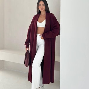 May include: A long, open-front burgundy cardigan worn over a white cropped tank top and white high-waisted jeans. The outfit includes white sneakers with dual hook-and-loop straps and a small, matching burgundy handbag held in one hand.